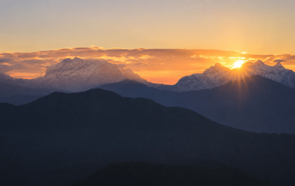 Sunrise view from Sarangkot, Pokhara, with golden sunlight rising behind the Annapurna mountain range and layered hills in the foreground.