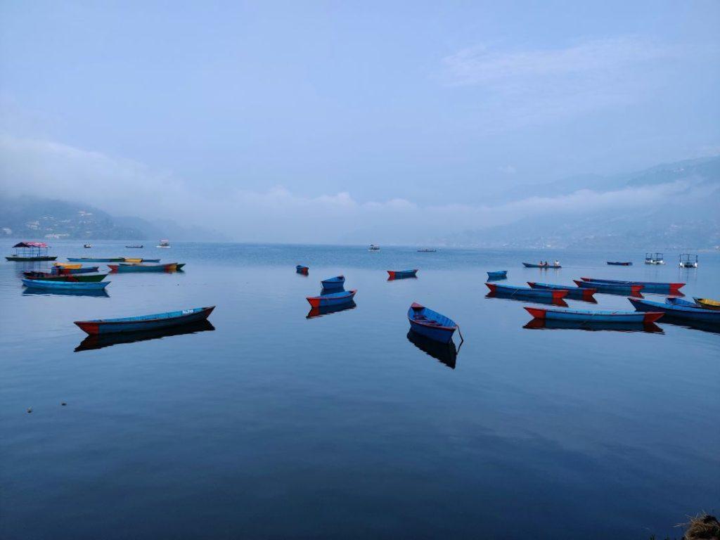 Colorful wooden boats on Phewa Lake at Lakeside Pokhara, Nepal, with calm water reflecting the surrounding hills and Annapurna Himalayan range in the background