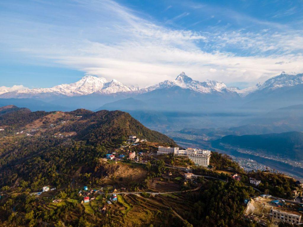 Drone view of Sarangkot viewpoint showing panoramic Himalayan mountain range, including Annapurna peaks, with mist-covered hills and Pokhara valley below.