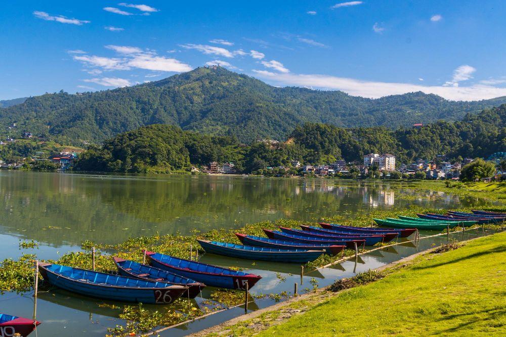 Colorful boats along the shore of Phewa Lake in Pokhara with green hills and clear blue sky, Nepal.